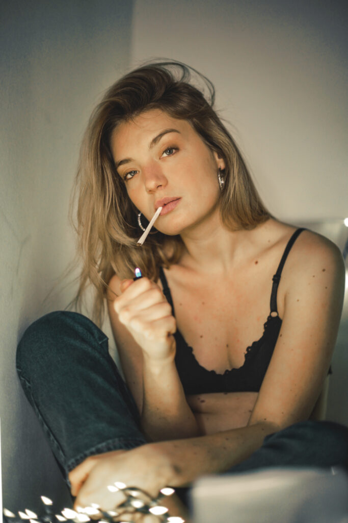 A shallow focus of a woman lighting a cigarette on her bed with light garlands on the background